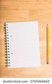 Spiral-bound lined notebook is lying open on wooden tabletop, with yellow pencil resting parallel. Stationery, writing, education, office supplies, creative process, organization, minimalistic