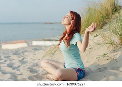 Spiritual Young Woman Sitting On A Beach Meditating Facing Into The Warmth Of The Sun With Eyes Closed And A Serene Expression