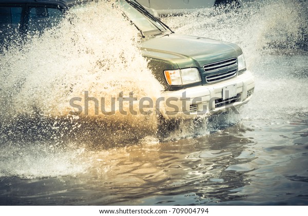 Splash by car as it goes through flood water after\
heavy rains of Harvey hurricane storm in Houston, Texas, US.\
Flooded city road big puddle spray from the wheels of SUV car\
roaring by. Vintage tone