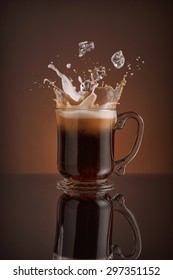 Splash Of  Ice Coffee Drink On A Brown Background. Refreshing Iced Cappuccino Liquid Drink Pouring Into A Mug, Cup With Ice Cubes. Cold Beverage Wave. Close-up Design Liquor Milk, Coffee And Ice.   