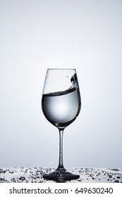 Splashing Transparent Water Wave In The Wine Glass While Standing On The Glass With Droplets Against Light Background.