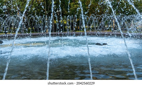 Splashing Water In The Fountain At Summer City Public Urban Park. Water Jets Abstract Image Close-up. Thin Jets Of Pure Clear Aqua Splashes Flying Upwards Under Pressure.