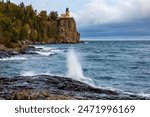 Split Rock lighthouse in Minnesota under grey sky.  Large rock bluff with blue and green water splash in foreground.  Motion blur action water against wet rocks.  