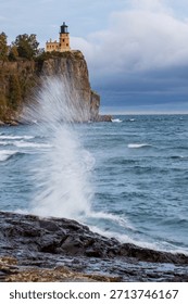 Split Rock lighthouse in Minnesota under grey sky.  Large rock bluff with blue and green water splash in foreground.  Motion blur action water against wet rocks.  