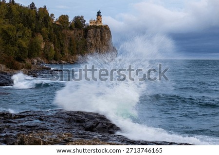 Split Rock lighthouse in Minnesota under grey sky.  Large rock bluff with blue and green water splash in foreground.  Motion blur action water against wet rocks.  