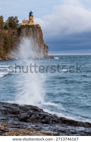 Split Rock lighthouse in Minnesota under grey sky.  Large rock bluff with blue and green water splash in foreground.  Motion blur action water against wet rocks.  