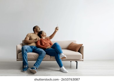 Split system. Cheerful young black couple with remote relaxing under air conditioner