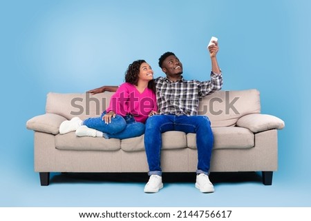Split system. Cheerful young black couple with remote relaxing under air conditioner, sitting on couch over blue studio background. Happy millennial spouses using climate control Stockfoto © 