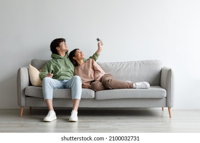 Split system. Happy young Asian couple with remote relaxing under air conditioner, sitting on couch at home, mockup for design. Happy millennial spouses using climate control indoors