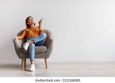 Split system. Happy young Asian woman with remote relaxing under air conditioner, sitting in armchair against white studio wall, mockup. Happy millennial lady using climate control