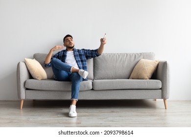 Split System. Portrait Of Middle Eastern man suffering from summer heat, sweaty guy holding remote control switching on ac air conditioner, sitting on couch in living room cooling himself waving hand - Shutterstock ID 2093798614