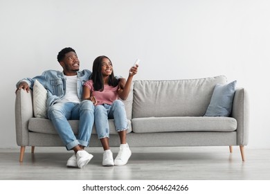 Split System. Satisfied African American woman holding remote control relaxing under the air conditioner, happy millennial black couple sitting on couch in living room, free copy space