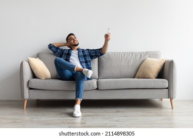 Split System. Satisfied Arab man holding remote control relaxing under the air conditioner, happy millennial guy sitting on couch in living room, adjusting temperature mode on ac, free copy space - Shutterstock ID 2114898305