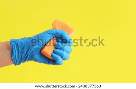 Sponge in hand, wet cleaning of the room, bright yellow background. A woman's hand in a latex blue glove with an orange sponge. Cleaning. Wash dishes
