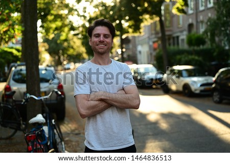 Spontaneous male master student smiling and standing with his arms crossed in a traditional street in Amsterdam, the Netherlands