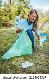 Spontaneous Photo Of A Cute Young Adult Blondie, Cleaning Up The Park On A Beautiful Sunny Day. She’s In A Squatting Position, Putting Garbagetrash In A Big Blue Bag 