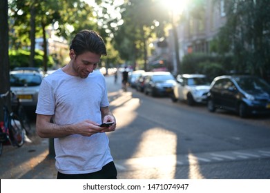 Spontaneous And Smiling Young Man Texting His Girlfriend On His Mobile Phone While Standing In An Old And Ambient Street