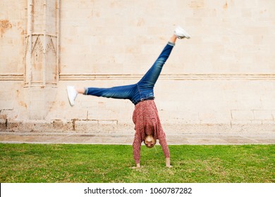 Spontaneous Young Woman In City Park Holiday, Doing Cartwheel On Grass In Front Of Stone Wall Texture Building, Outdoors. Teenager Celebrating Dynamic Energy Fun, Travel Recreation Leisure Lifestyle.