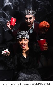 Spooky African American Woman In Werewolf Mask Holding Toy Hand Near Scary Man With Carved Pumpkin On Black