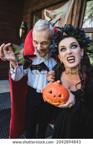 spooky couple in halloween vampires costumes grimacing with carved pumpkin and paper cut bat