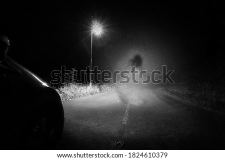 A spooky ghost of a woman in the road below a single street light in the countryside. Lit up by car headlights at night. With a grunge, vintage, old edit