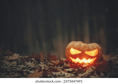 Spooky glowing Jack o lantern on autumn leaves in moody dark forest. Happy Halloween! Scary atmospheric halloween carved pumpkin in evening fall woods. Boo! Horror time. Copy space