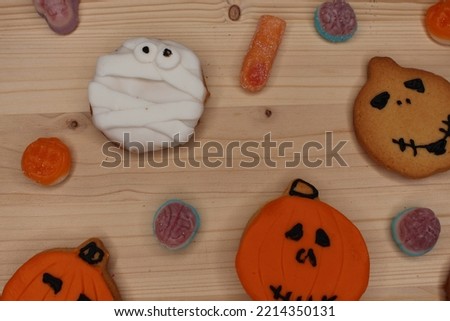 Spooky Halloween cookies on a light wood table. White and orange fondant and icing. There are sweets and candies decorating the background. Wallpaper of a party, celebration and festivity.