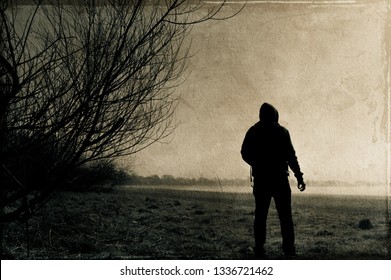 A spooky lone hooded figure. Standing on the edge of a field on a cold, misty winters night. With a sepia, grunge, vintage edit.