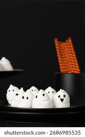 Spooky marshmallow ghosts with hand-drawn faces on a black plate and orange patterned straws in the background. Halloween party concept
