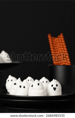 Spooky marshmallow ghosts with hand-drawn faces on a black plate and orange patterned straws in the background. Halloween party concept