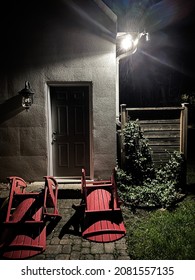 Spooky Shadows Cast At Night In A Residential Backyard. Three Adirondack Chairs Are Tipped Over And Laying On The Sides On A Stone Patio.