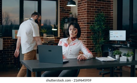 Spooky Zombie Sitting At Desk While Waving At Video Conference Call On Laptop. Creepy Brain-eating Monster With Bloody And Deep Wounds In Office Workspace Having An Online Conversation On Computer.