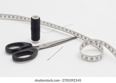 A spool of black sewing thread with a needle, scissors and a clothes measuring tape next to it isolated on a white background.