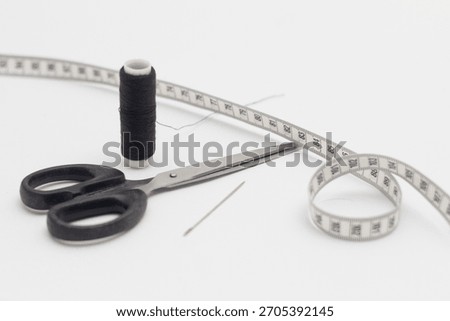 A spool of black sewing thread with a needle, scissors and a clothes measuring tape next to it isolated on a white background.