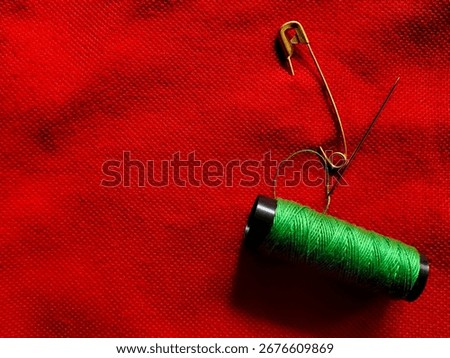A spool of green thread lies on a textured red fabric, accompanied by a gold safety pin and a steel sewing needle. 