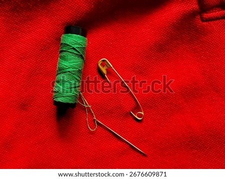 A spool of green thread lies on a textured red fabric, accompanied by a gold safety pin and a steel sewing needle. 