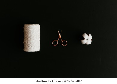 A Spool Of White Cotton Rope, A Rose Gold Scissors And A Cotton Flower Head On Black Background.