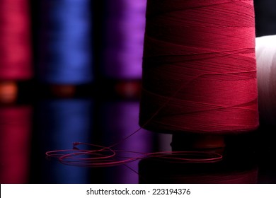 Spools Of Colored Cotton Thread, Ordered Composition, Warm Colors, Red Spool In The Foreground With The Red Wire Coiled In The Form Of  Black Table Reflection, Sprockets Into The Background Blurred