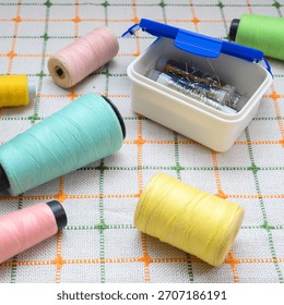 spools of multi colored threads and an open plastic container with gold and silver wire, casually scattered on a table covered with a white green checkered tablecloth, for sewing