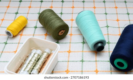spools of sewing threads in various colors and a plastic container with gold and silver wire, laid out on a table surface with a white green checkered tablecloth