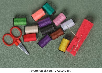 Spools thread in different colors, scissors and sewing needles on green leather background