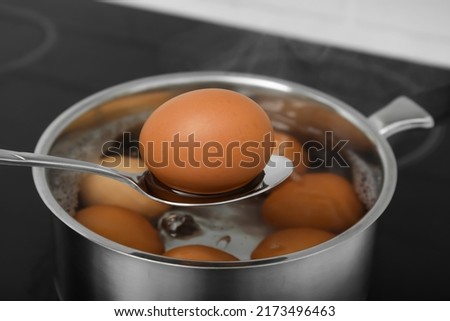 Spoon with boiled egg above saucepan on electric stove, closeup
