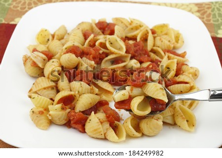 Spoon lifting out a taste of small conchigliette pasta shells prepared with diced tomatoes, many peeled garlic cloves, parsley, basil and oregano served on a white plate with a colorful background.