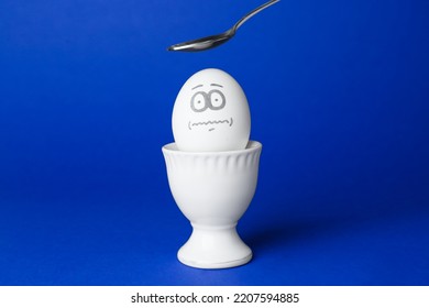 Spoon Over Egg With Drawn Frightened Face Among Others In Cup On Blue Background