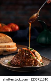 Spoon Pouring  Gravy On To A Pot Roast With Mashed Potatoes, Sweet Peas And Shredded Carrots On Dark Rustic Background With Bread And Tomatoes.