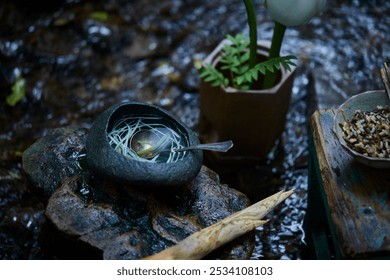 A spoon rests in a dark stone bowl with delicate noodles. Nearby, a bamboo shoot and a leafy plant in a vase create an earthy setting. - Powered by Shutterstock