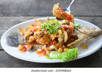 Spoon To Scoop Up A Piece Of Fried Fish From Plate Of Thai Food Name Deep-Fried Snapper With Three-Flavoured Sauce. Deep Fried Fish With Chili Sauce Served In A White Plate On A Blurred Background. 