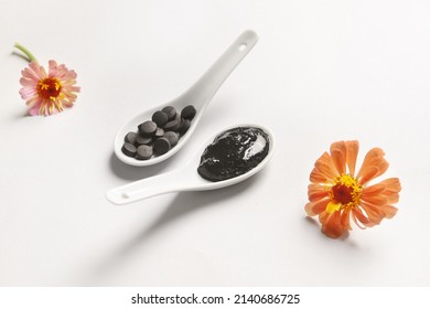 Spoons With Activated Carbon Mask, Pills And Flowers On White Background