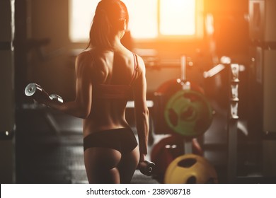 Sport. Athletic fitness woman pumping up muscles with dumbbells. Brunette sexy fitness girl in pink sport wear with perfect body in the gym posing before training set. Fitness woman in the gym. 