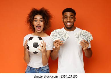 Sport bet excitement and family lifestyle. Excited african-american couple enjoying their win, standing with soccer ball and lots of money, orange background - Shutterstock ID 1354127150